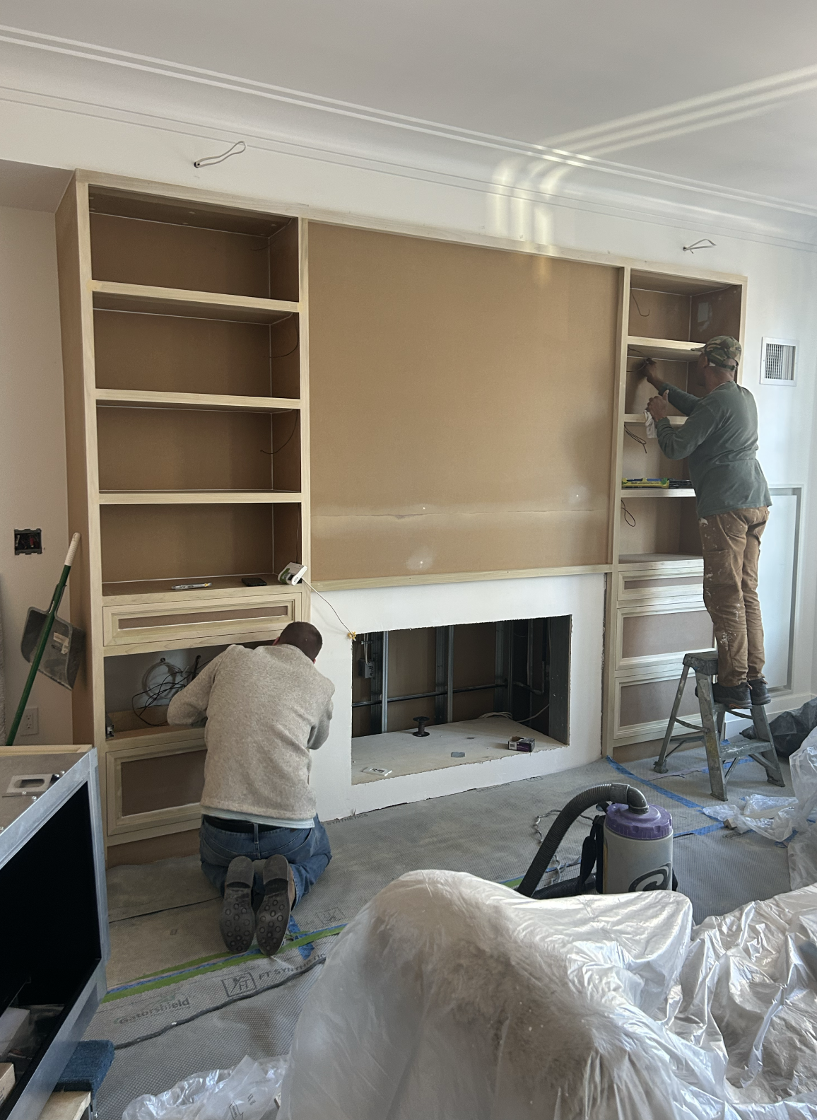 Two workers installing built-in shelves around a fireplace in a living room.